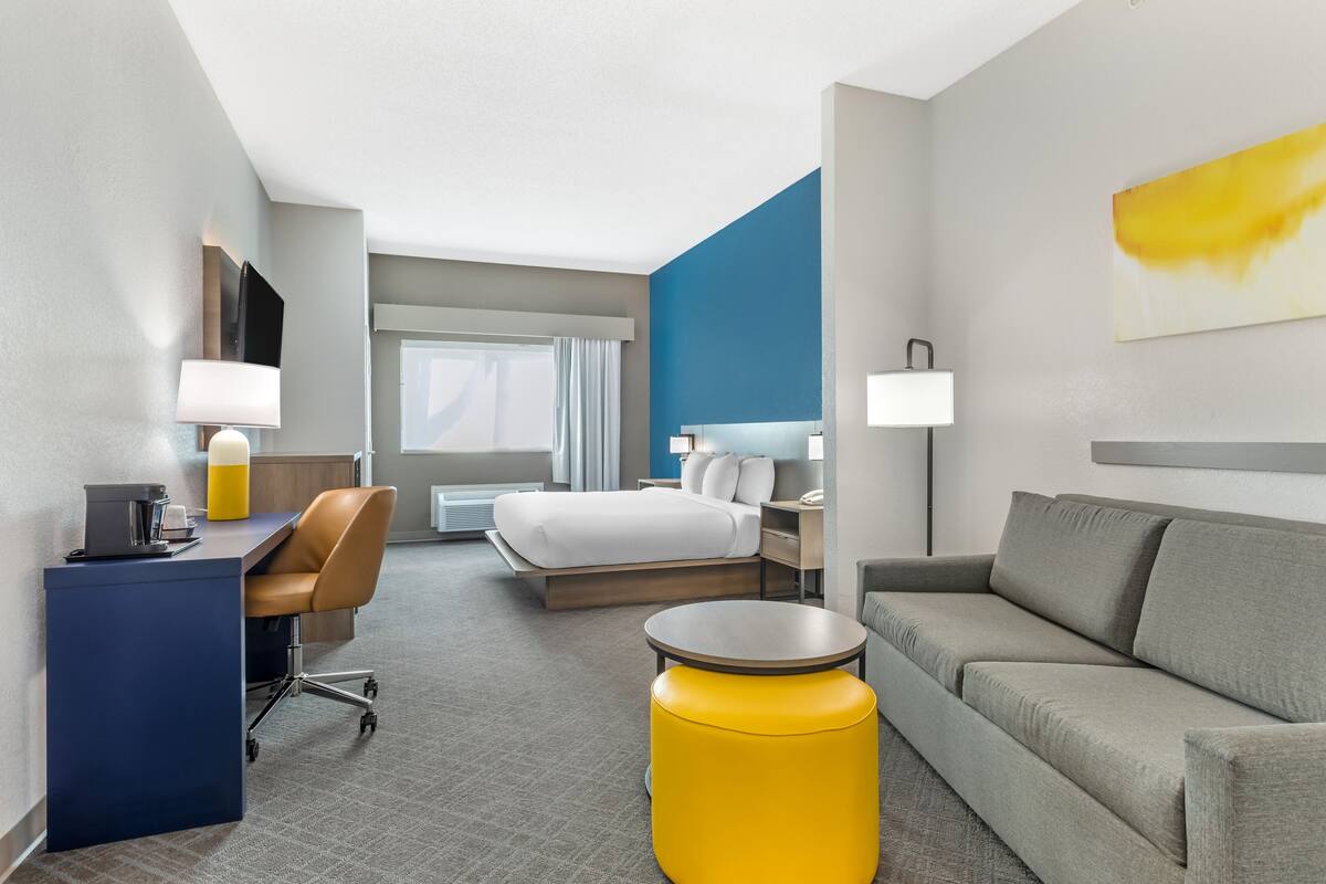 A modern hotel room with a bed, a desk, a sofa, and a yellow ottoman.