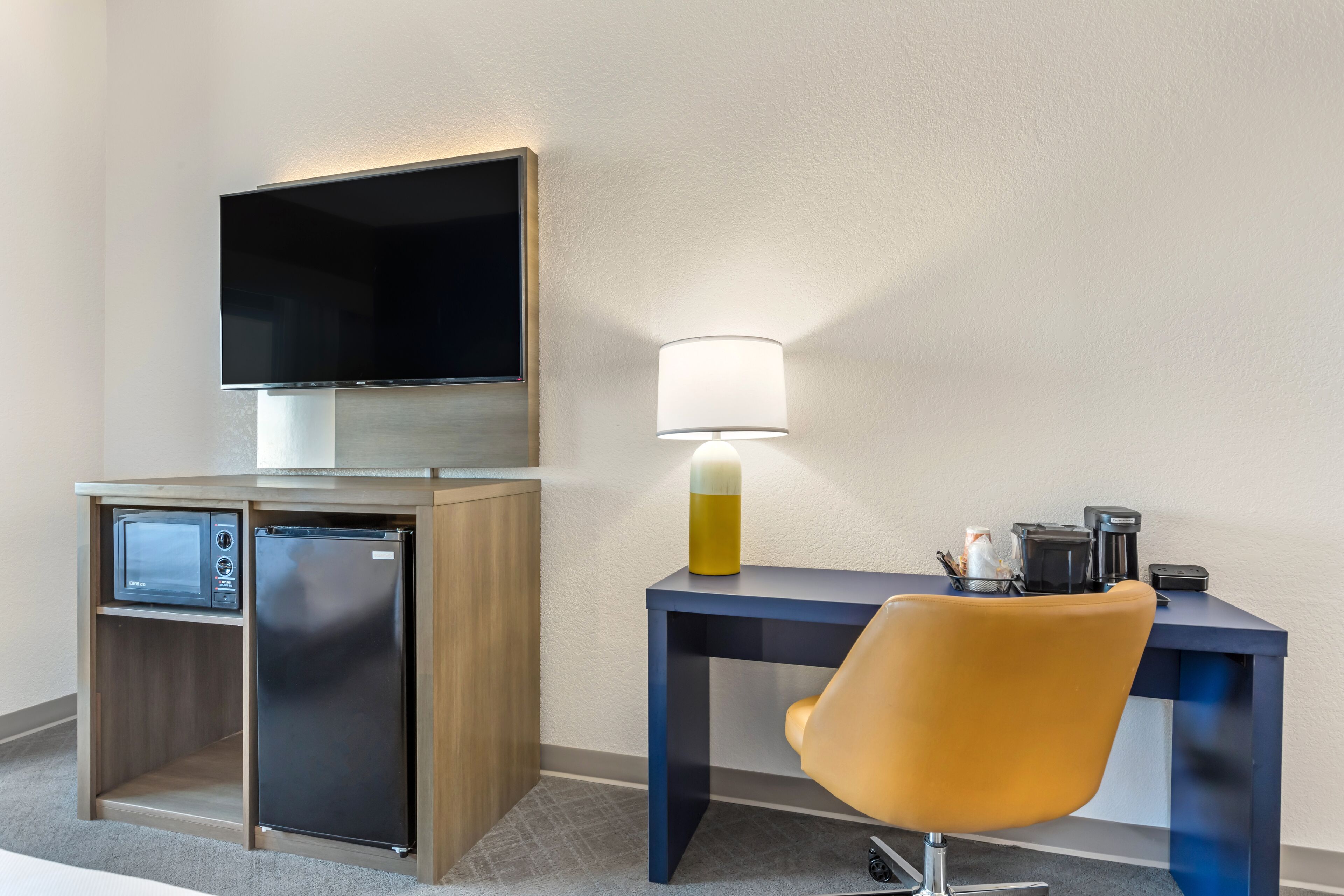 Habitación de hotel compacta con televisión de pantalla plana en la pared, un refrigerador pequeño, un escritorio con lámpara y una sola silla.