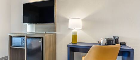 Habitación de hotel compacta con televisión de pantalla plana en la pared, un refrigerador pequeño, un escritorio con lámpara y una sola silla.