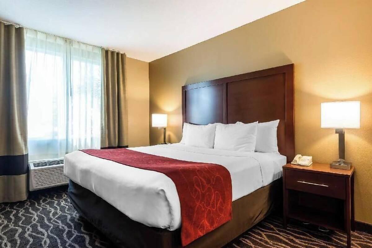 A hotel room with a large bed, two bedside lamps, a red throw blanket, a wooden headboard, a nightstand with a phone, and a window with curtains.
