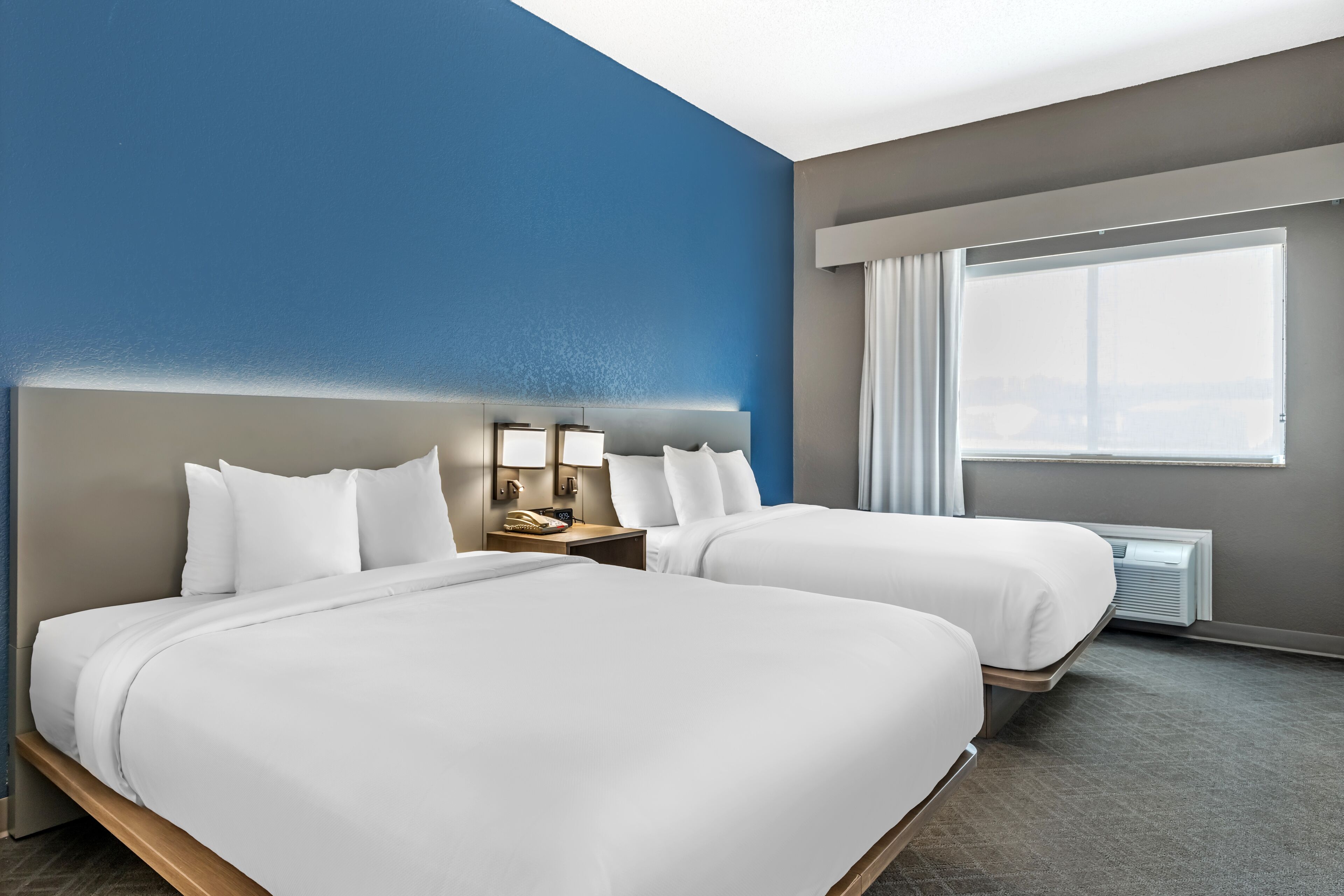 A hotel room with two beds, a large window with curtains, a nightstand, and a wall-mounted lamp.