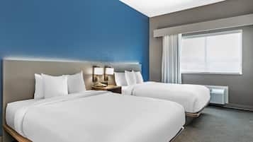 A hotel room with two beds, a large window with curtains, a nightstand, and a wall-mounted lamp.