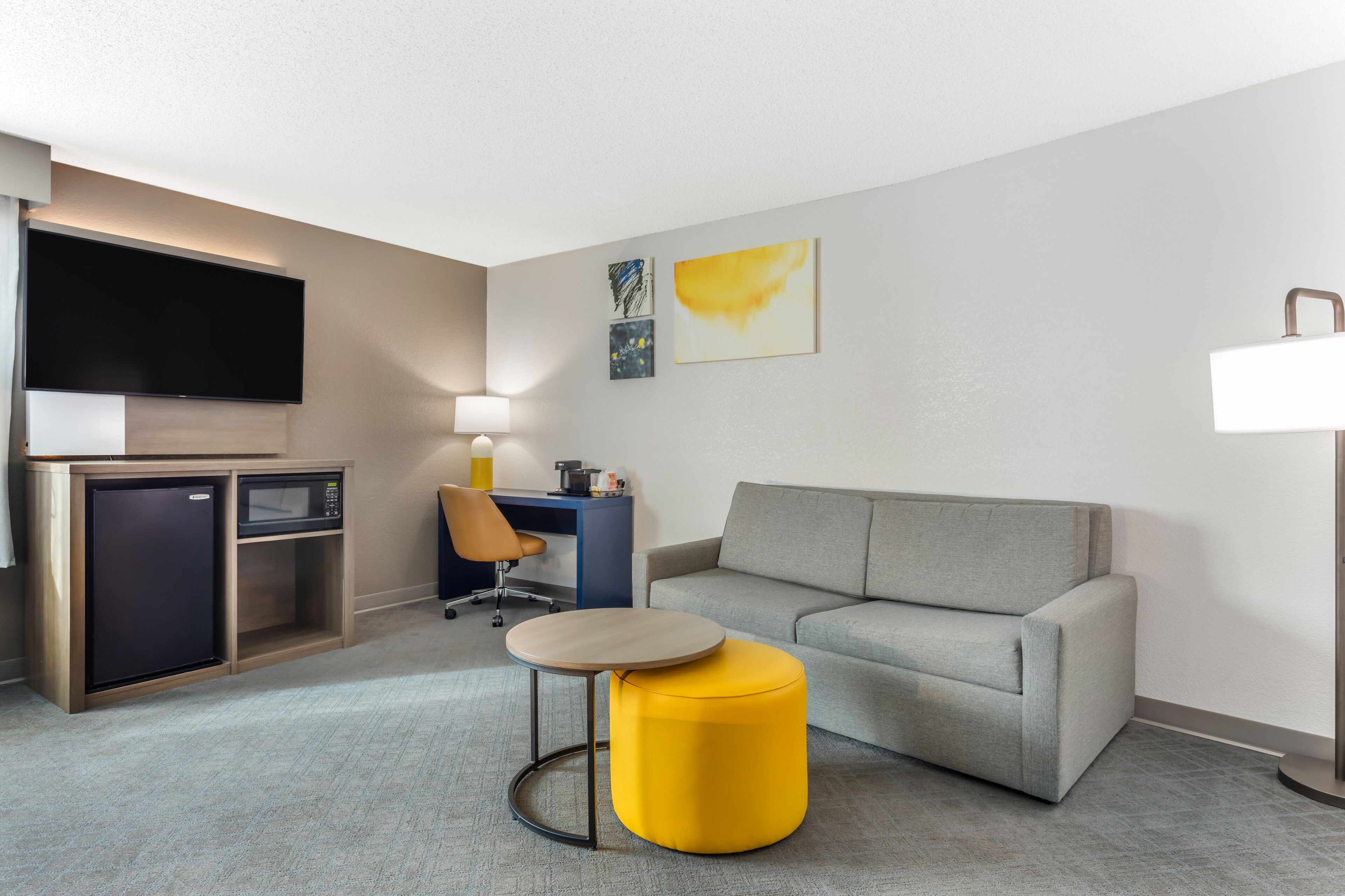 A modern hotel room with a grey sofa, a yellow ottoman, a desk with a chair, a flat-screen TV, a mini-fridge, and a microwave.
