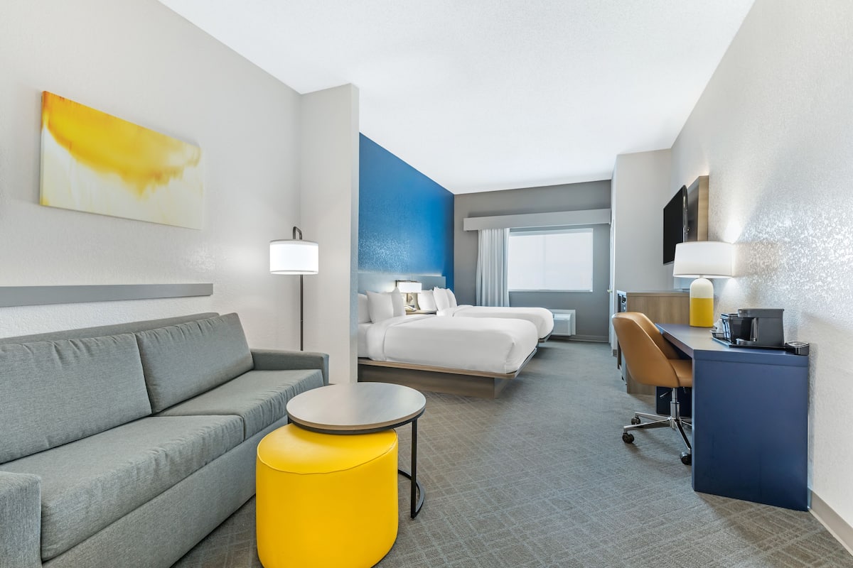 A hotel room with a grey sofa, a yellow ottoman, a bed with white linens, a desk with a chair, and a large window with curtains.