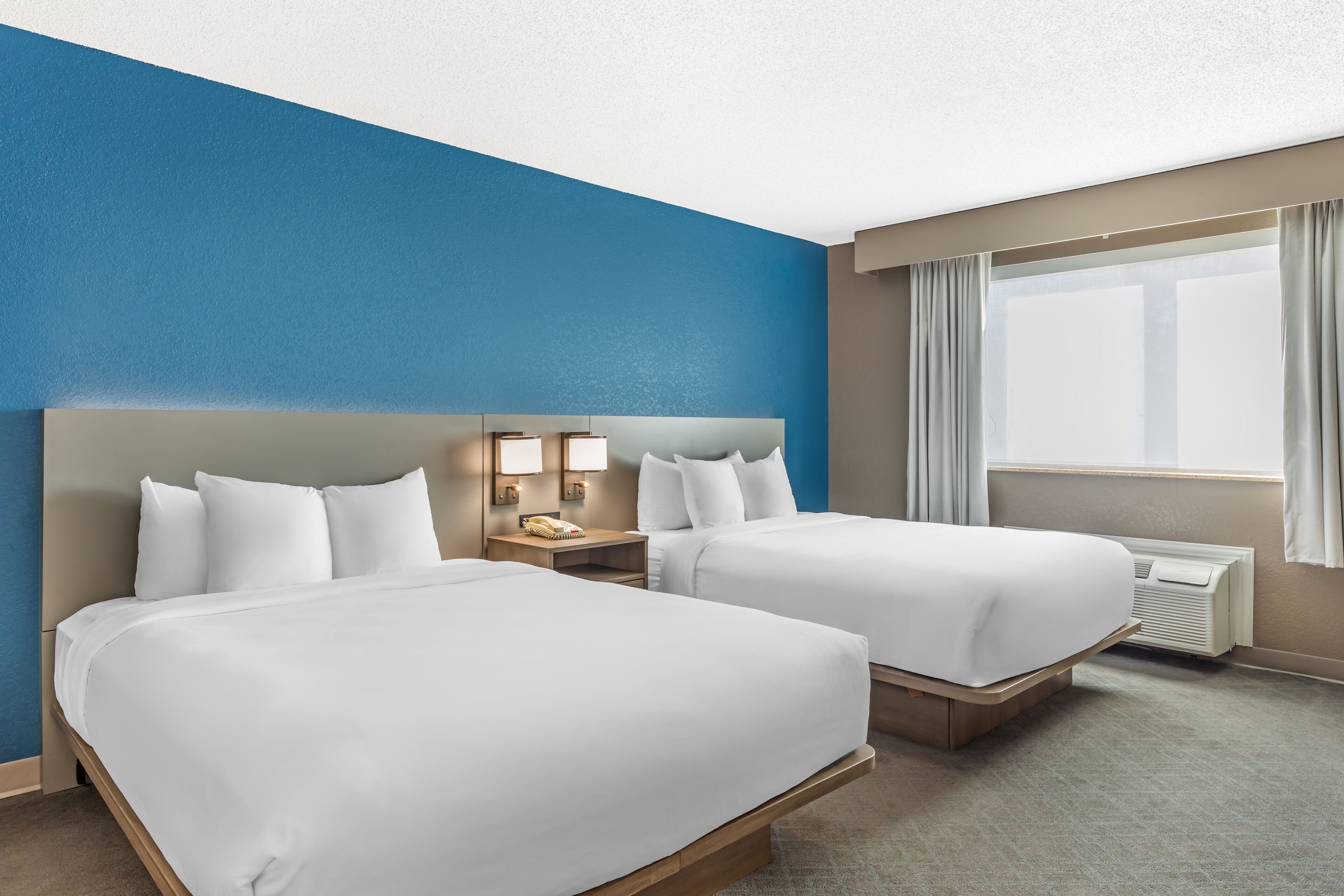 A hotel room with two beds, a nightstand with a lamp, a window with curtains, and a wall-mounted air conditioning unit.
