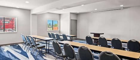 Un salón de conferencias con una larga mesa y sillas, una pantalla de proyector y un ventanal con vistas a edificios y un vehículo rojo.