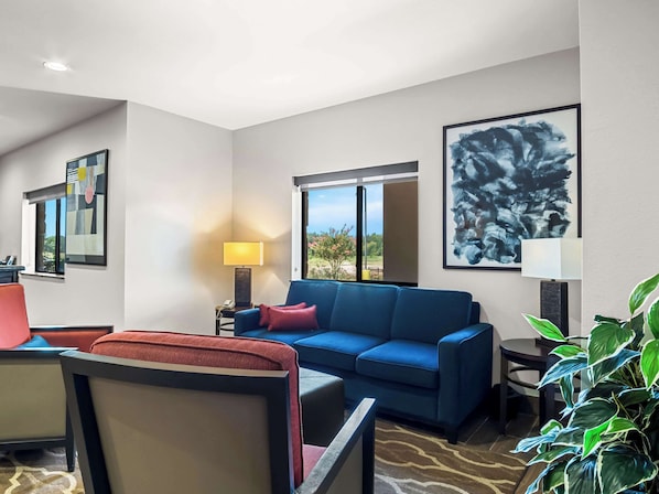 A hotel room with a blue sofa, a small table with a lamp, a large abstract painting, and a potted plant. - Comfort Suites Lindale - Tyler North (Lindale)