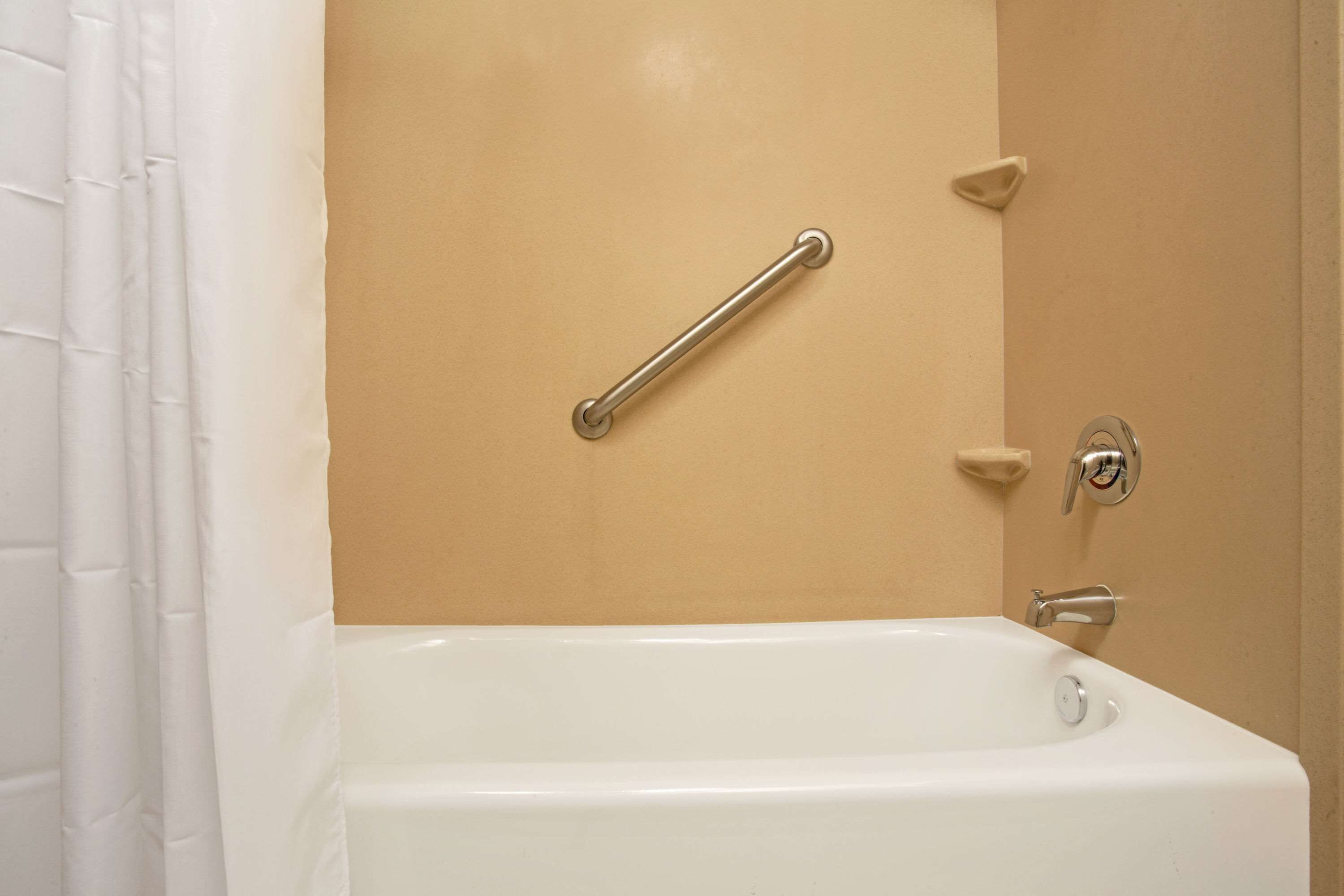 A bathroom with a white bathtub, a beige wall, and a grab bar installed on the wall.