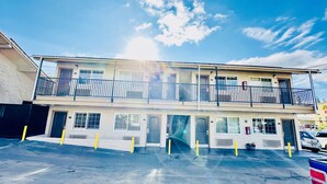 Un motel de dos pisos con balcón en el nivel superior, estacionamiento al frente y cielo despejado.