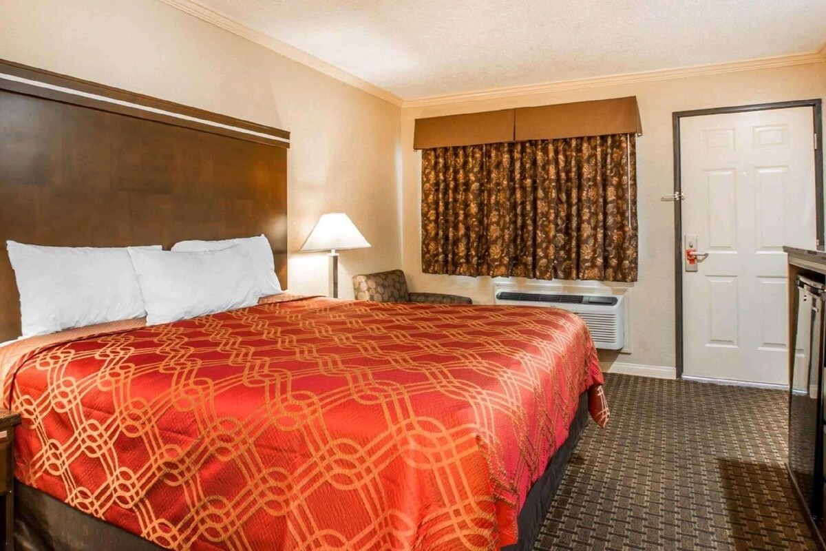 Une chambre d’hôtel avec un grand lit, un bureau, une chaise, une lampe, une fenêtre avec des rideaux et une porte.