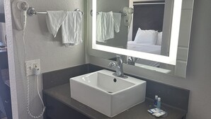 Un baño con lavamanos blanco, un espejo con iluminación integrada, un secador de pelo y un perchero para toallas.