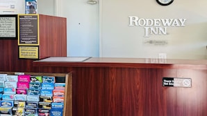 Mostrador de recepción en Rodeway Inn, con un reloj, un cartel que dice “Gracias por no fumar” y una exhibición de carteles de eventos locales.