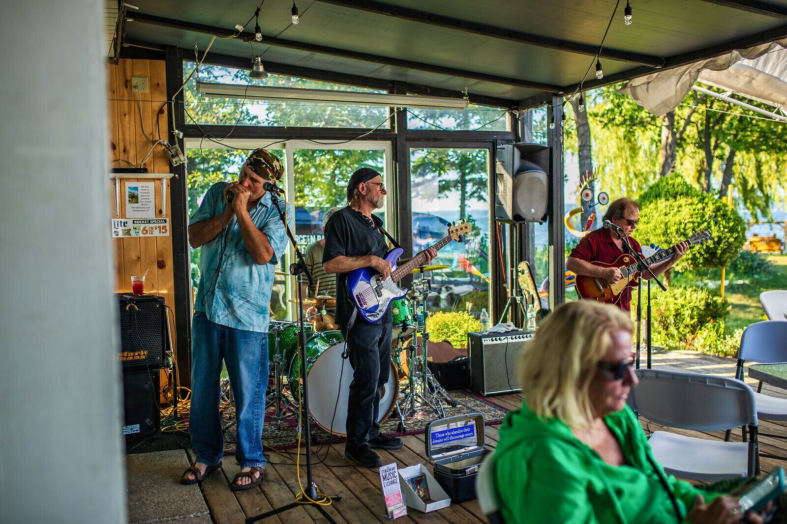 Een band treedt op in een overdekte buitenruimte met een houten vloer, een drumstel en een gitarist.