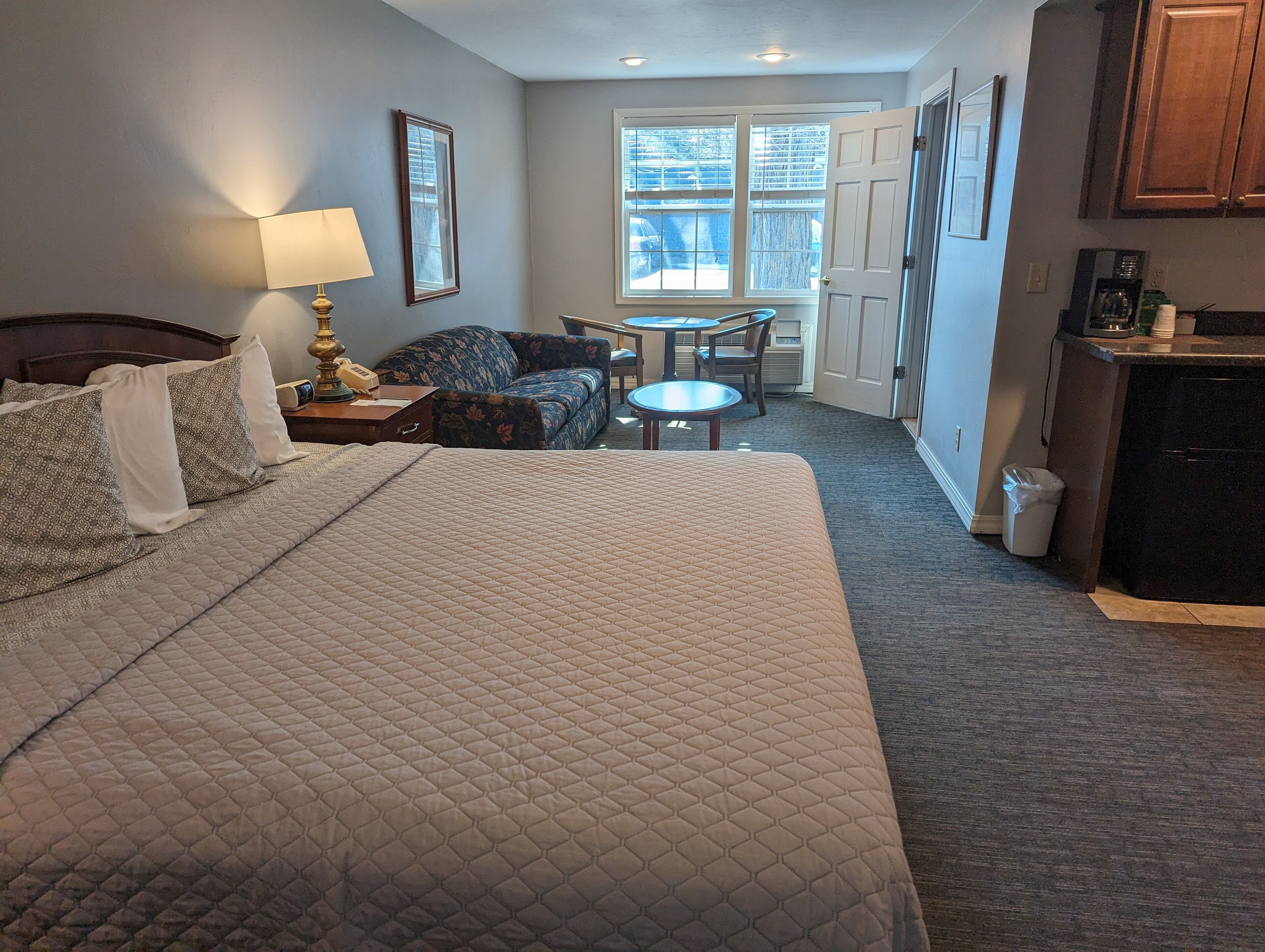 Habitación de hotel con una cama grande, un sofá, una mesita y una pequeña cocina.