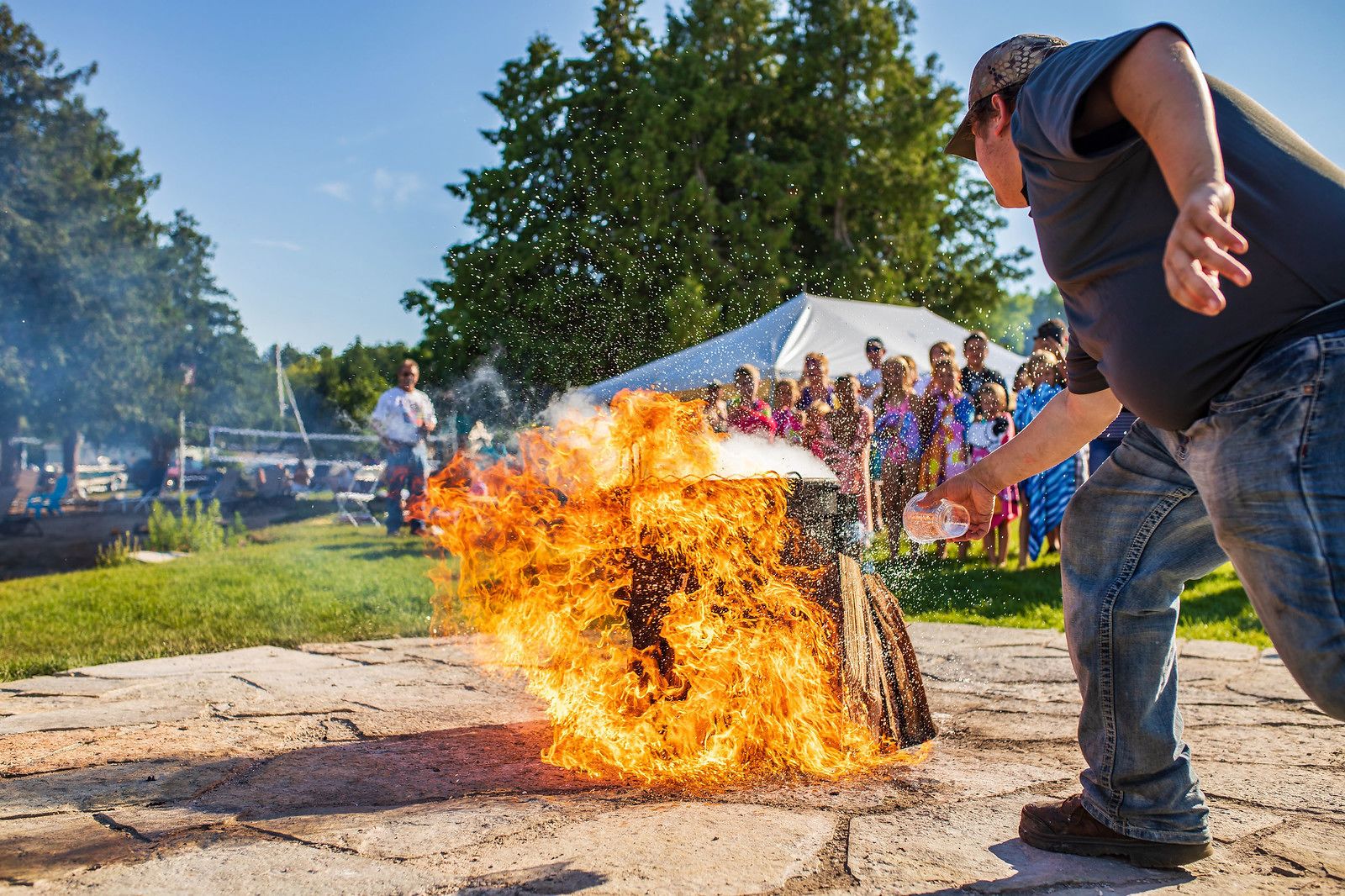 A person throwing a bottle of liquid onto a fire, causing a large flame to erupt.