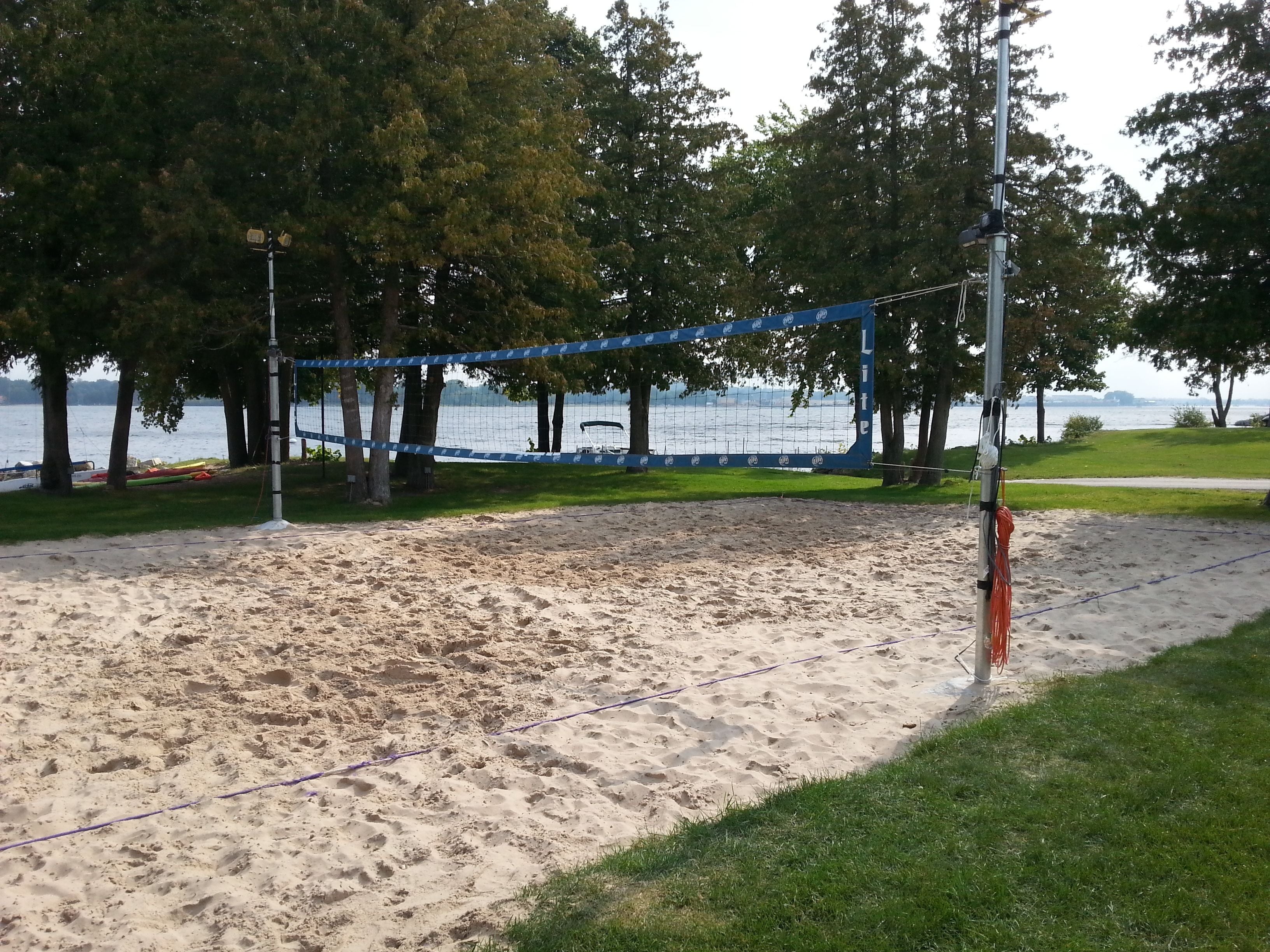 A sandy beach volleyball court with a net, surrounded by trees and a body of water in the background.