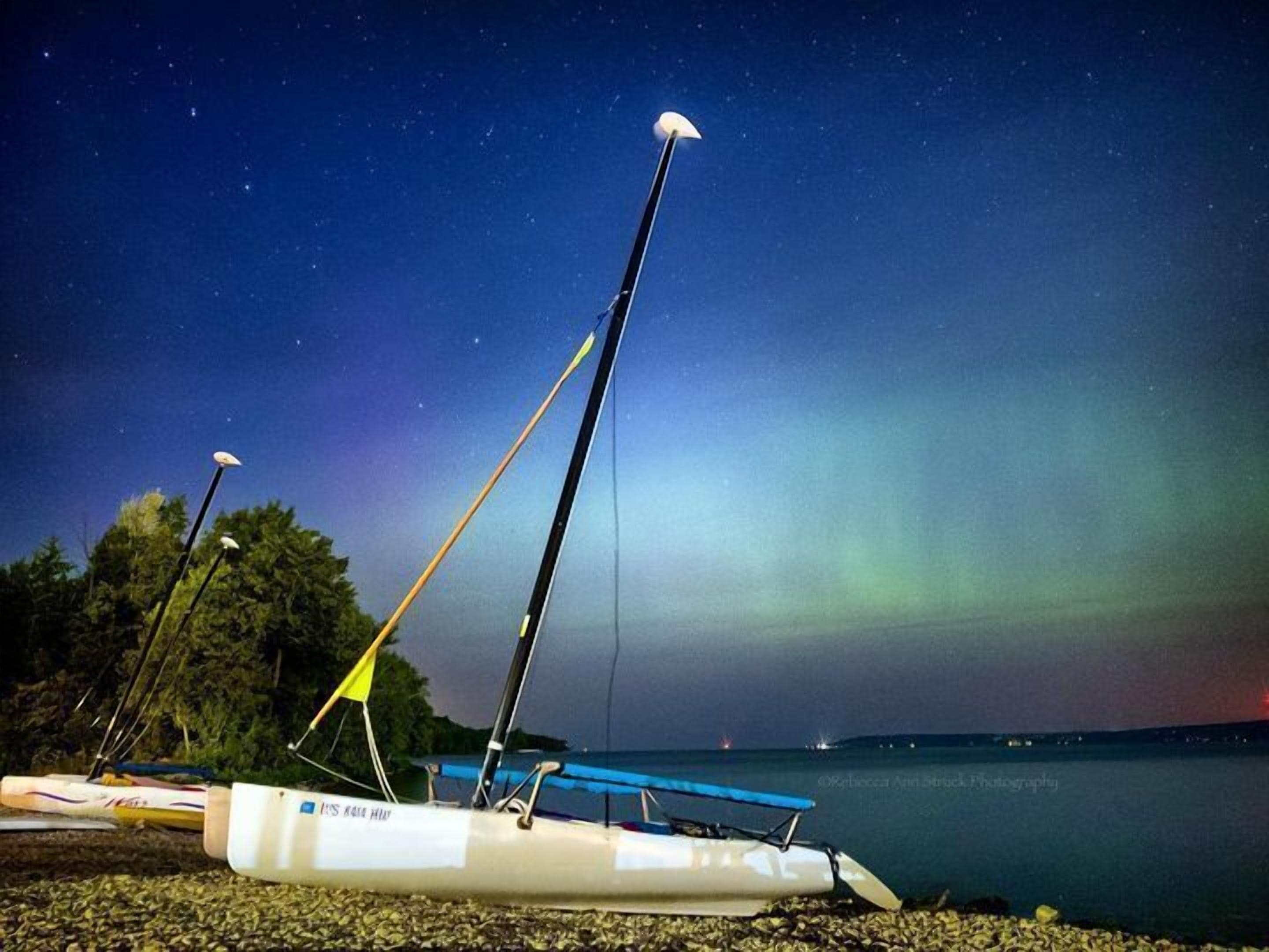 A sailboat with a white hull and a blue canopy, docked on a pebbled shore with trees in the background and a starry night sky.