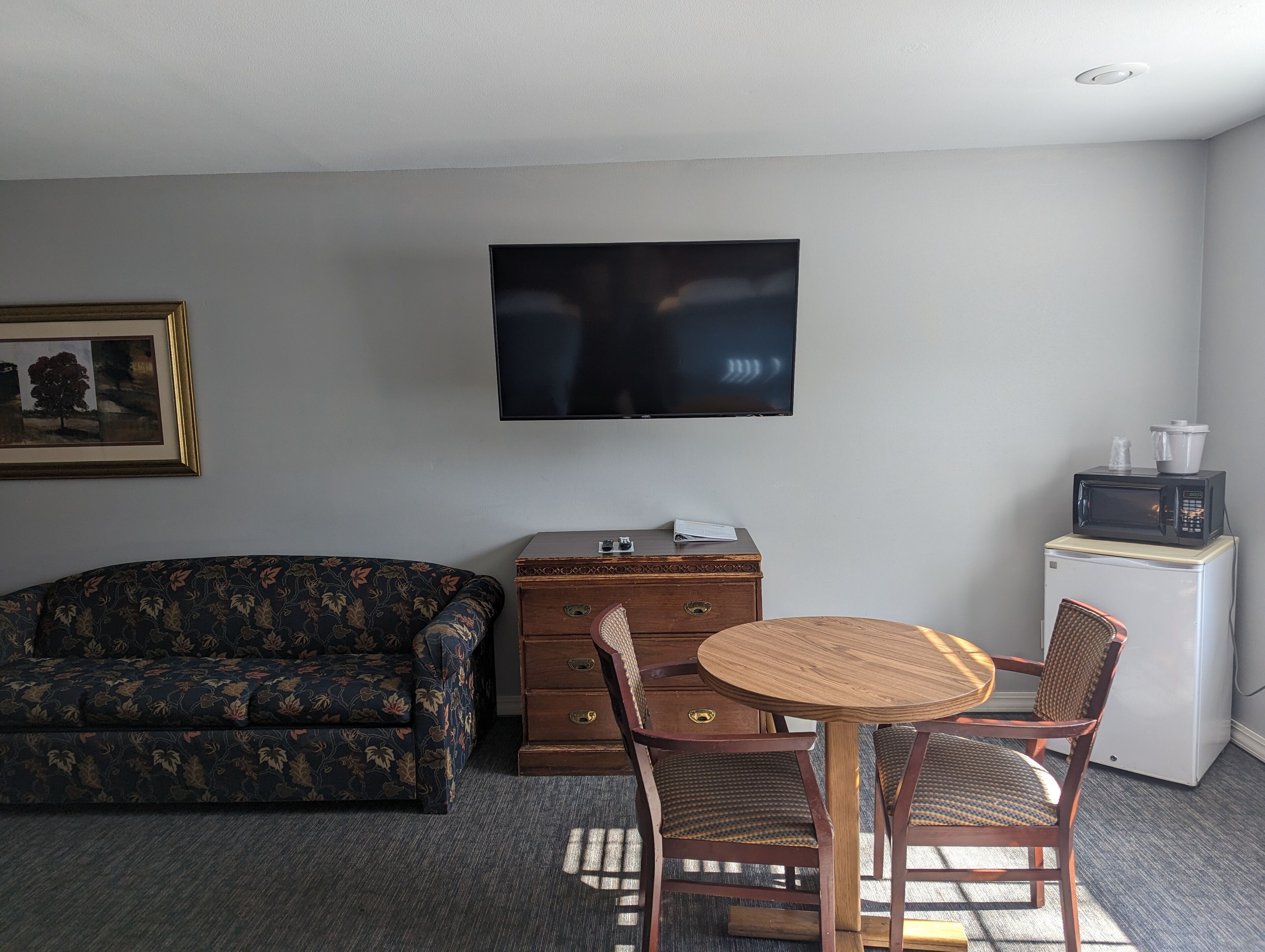 A room with a sofa, a wooden chest of drawers, a small refrigerator, a microwave, a television, and a round wooden table with two chairs.