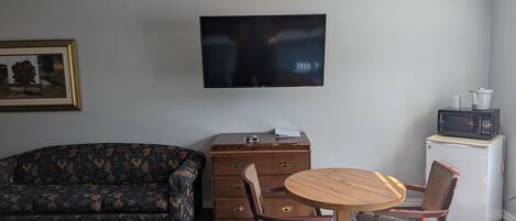 A room with a sofa, a wooden chest of drawers, a small refrigerator, a microwave, a television, and a round wooden table with two chairs.