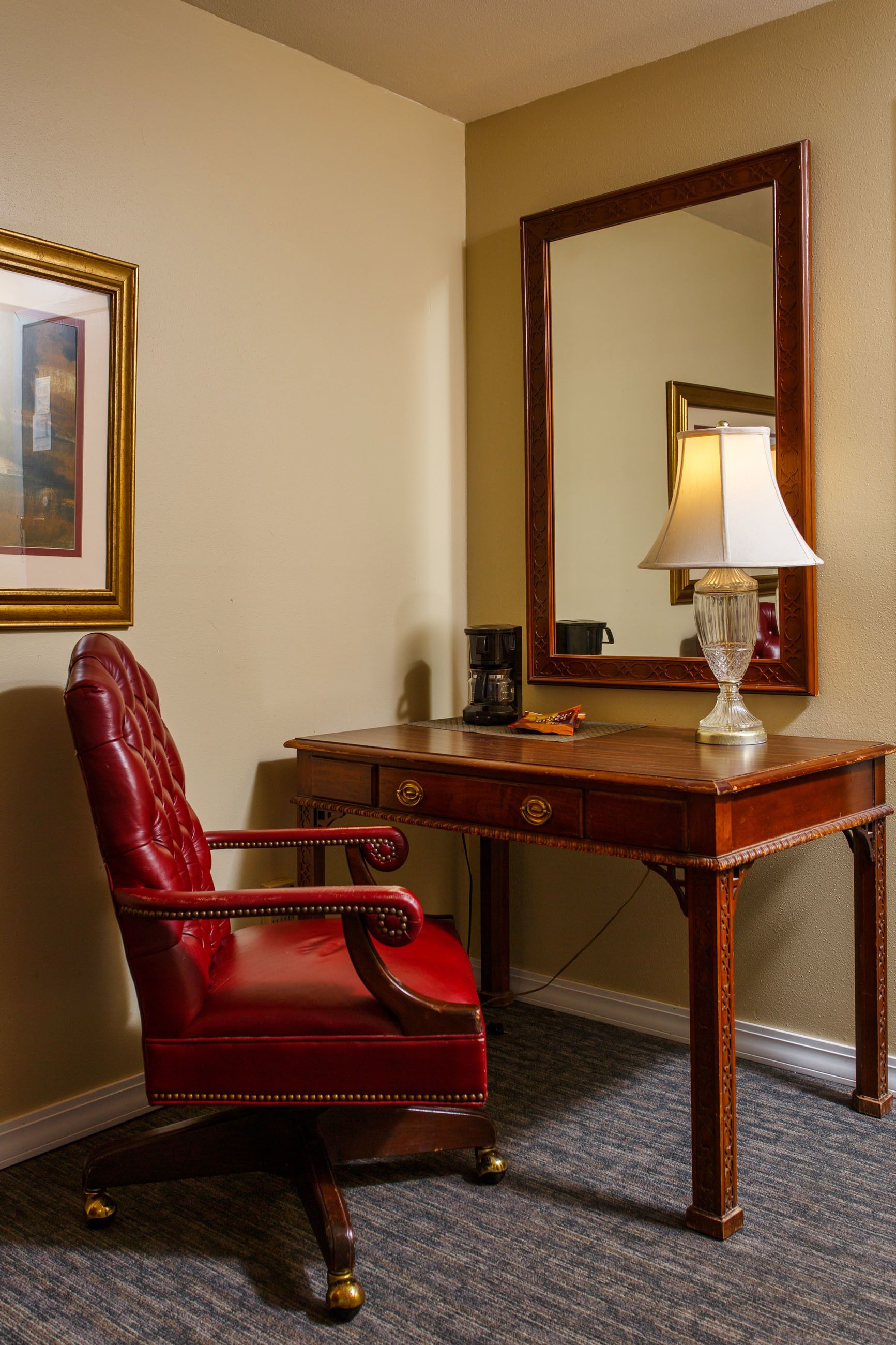 A room with a red leather chair, a wooden desk, a lamp, a framed picture, and a large mirror.