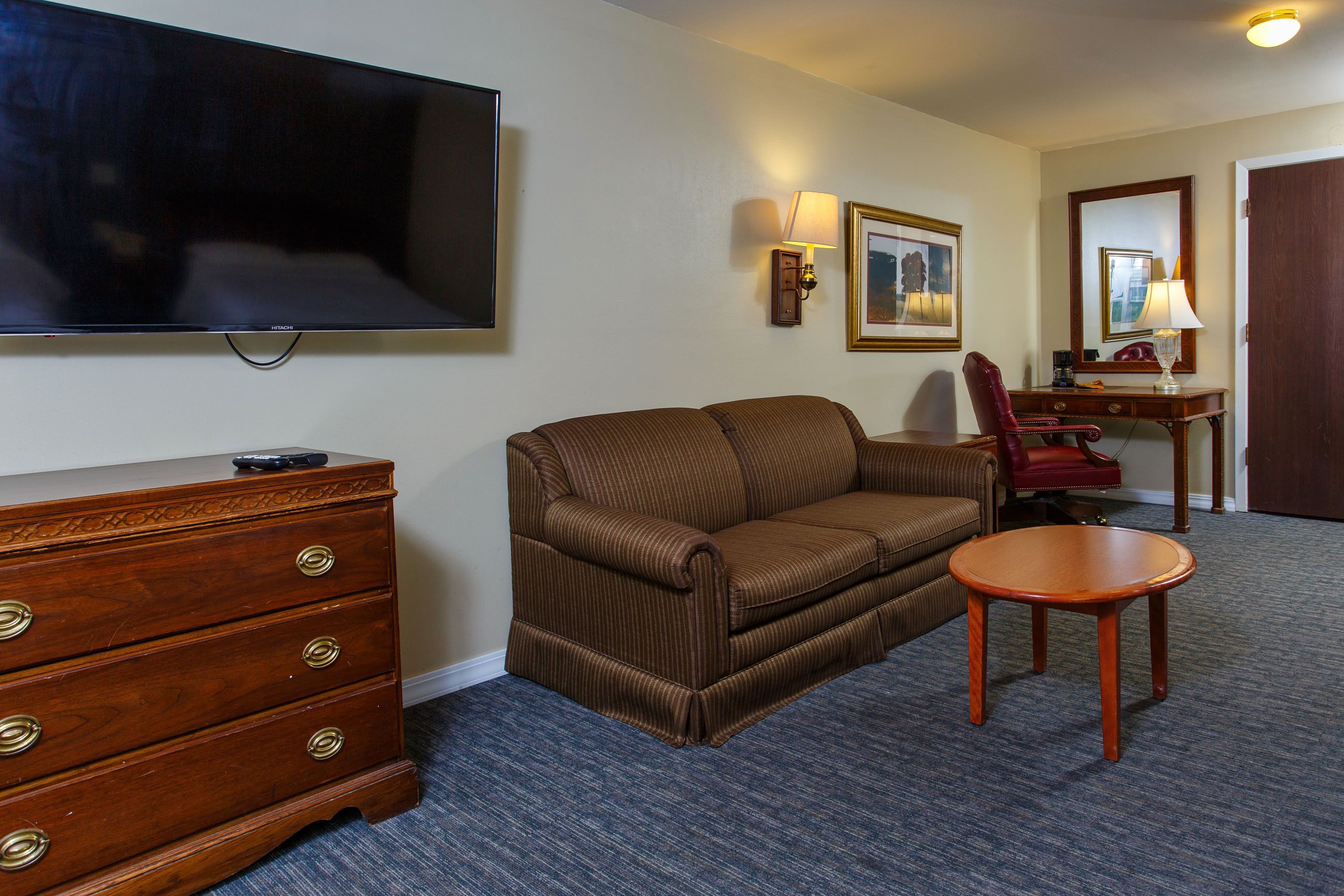 A hotel room with a brown sofa, a wooden dresser, a flat-screen TV, a desk with a lamp, and a chair.