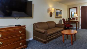 A hotel room with a brown sofa, a wooden dresser, a flat-screen TV, a desk with a lamp, and a chair.