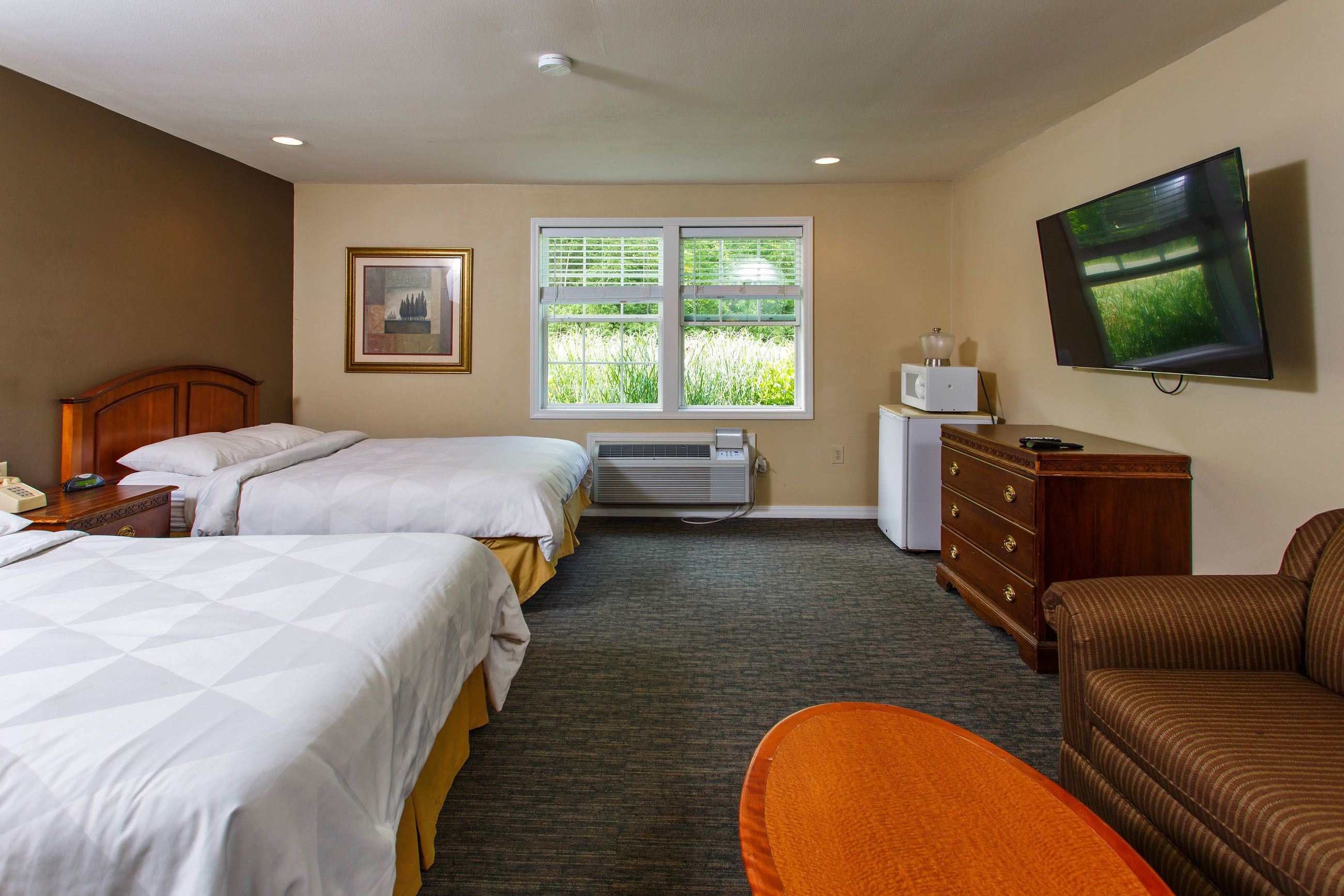 A hotel room with two beds, a TV, a dresser, and a window with a view of greenery.