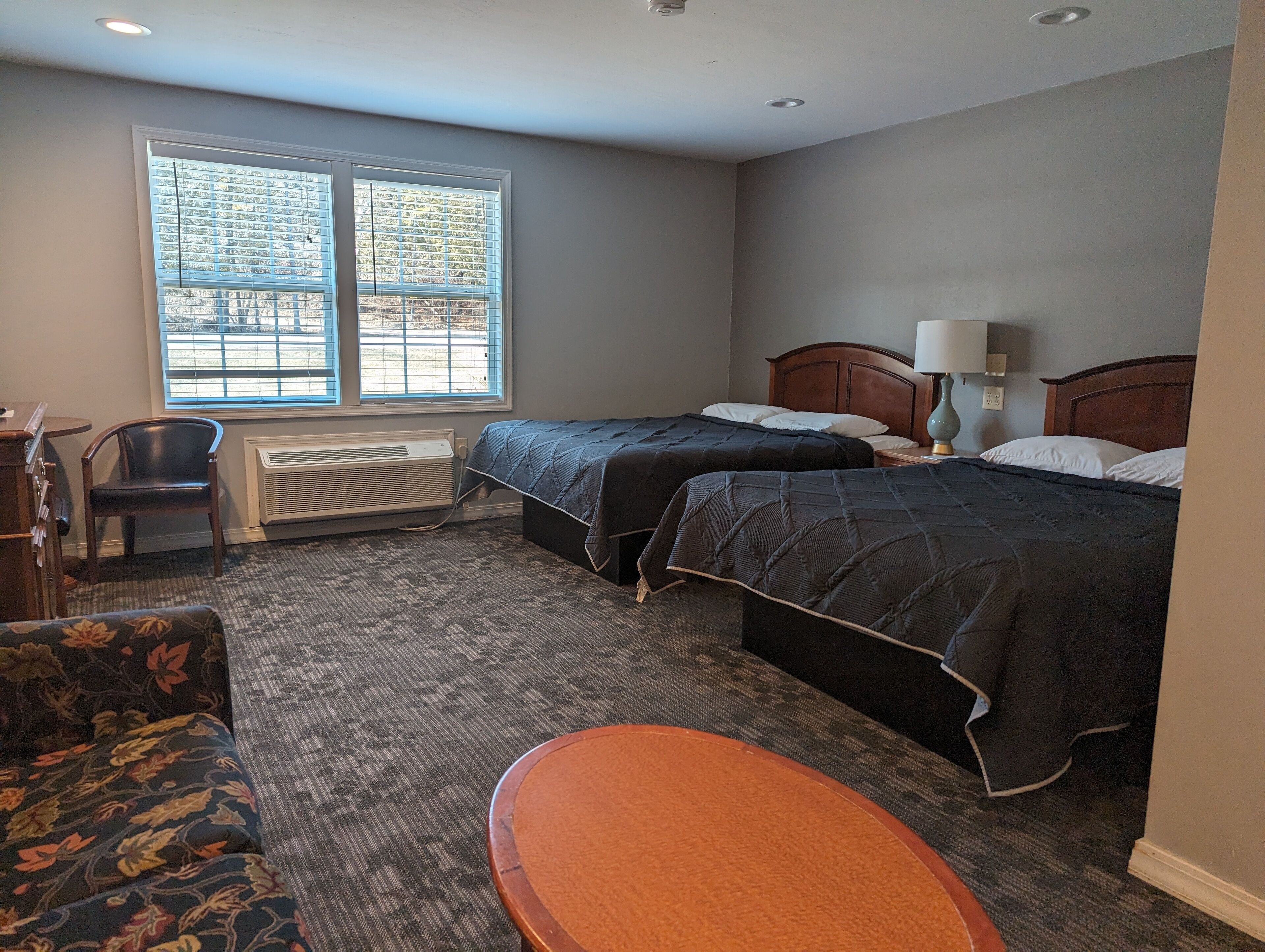 A hotel room with two beds, a desk, a chair, and a large window with blinds.