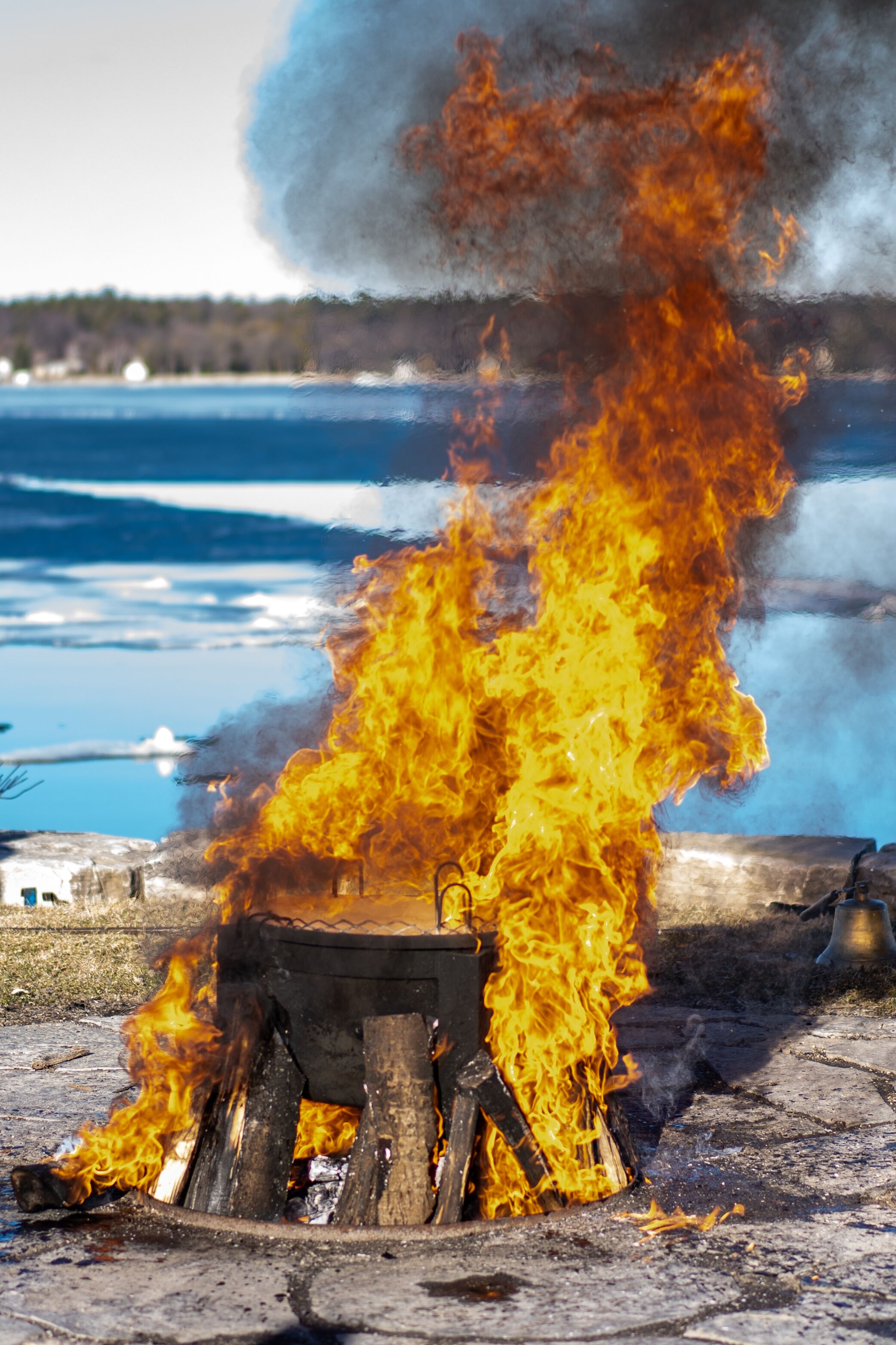 Un feu de camp avec des bûches et une marmite, entouré d’un paysage enneigé et d’une étendue d’eau.