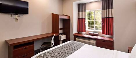 Habitación de hotel con cama, escritorio, silla, televisor montado en la pared y ventana con cortinas.