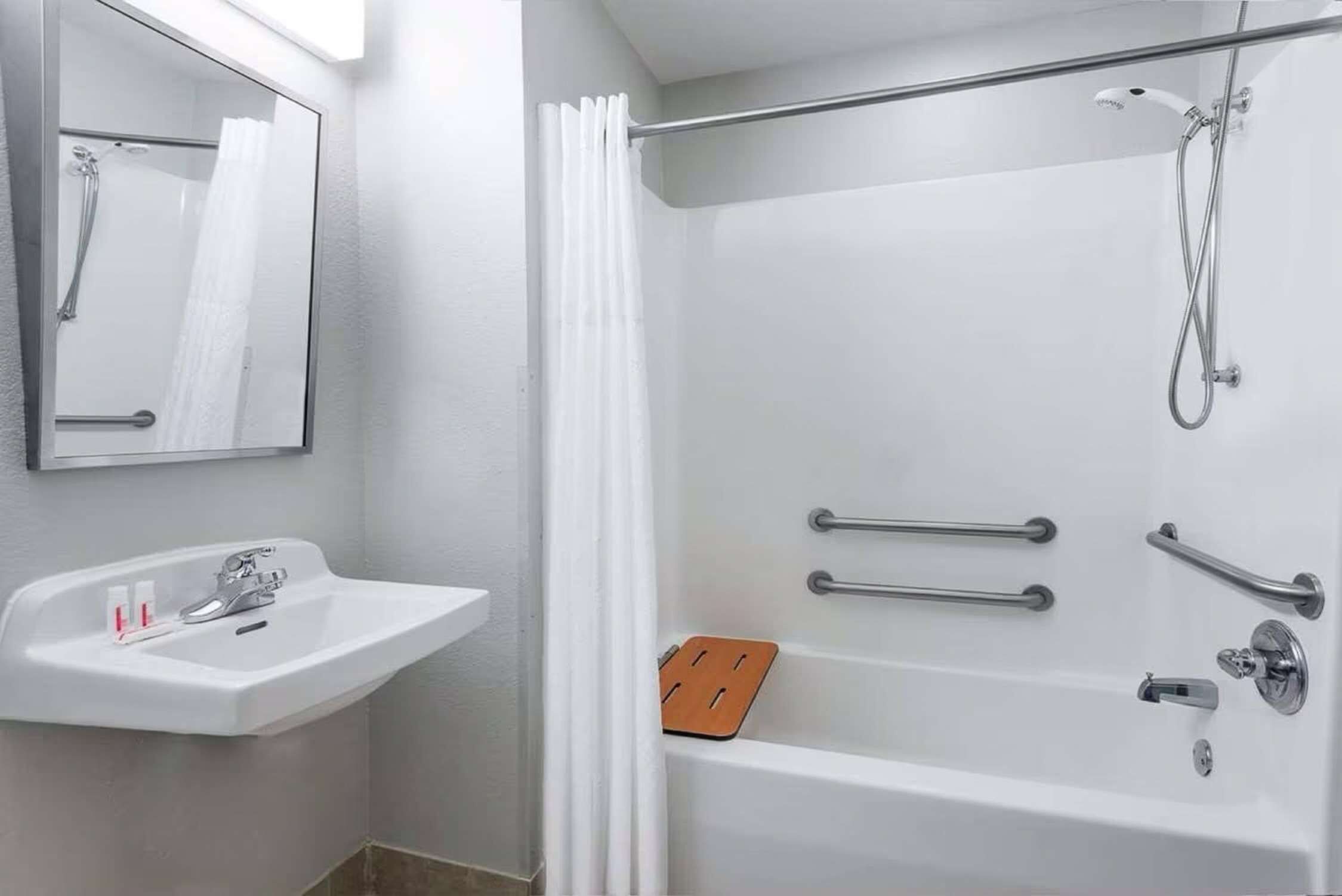 Un baño moderno con lavamanos blanco, espejo, ducha y barras de apoyo.