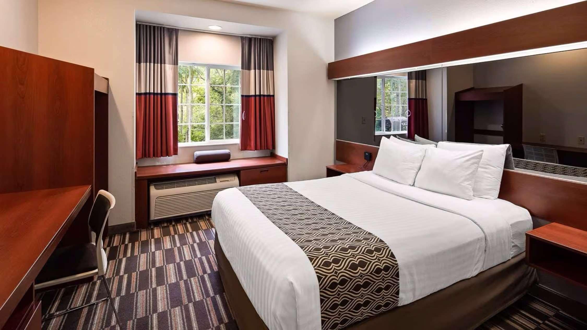 A hotel room with a large bed, a desk, a chair, a window with curtains, and a mirror.