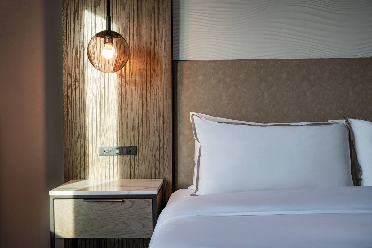 Una cama bien hecha con sábanas blancas, una mesita de noche de madera con cajón y una lámpara de pared.