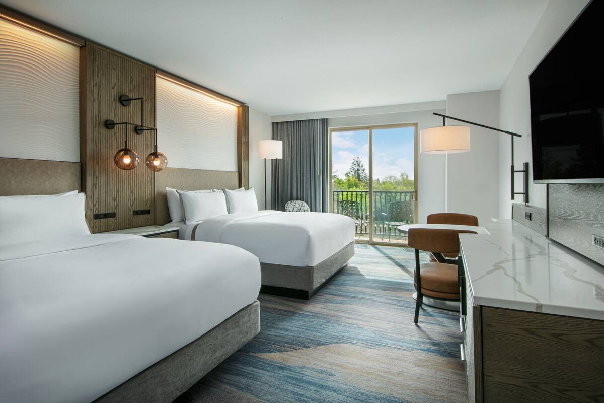 Habitación de hotel con dos camas, un escritorio, un televisor y un balcón con vistas.