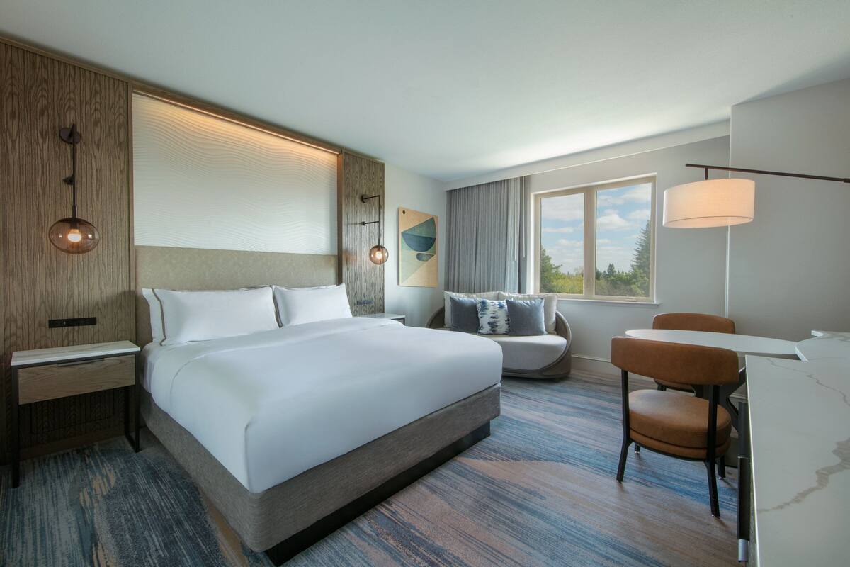 Una habitación de hotel moderna con una cama grande, un escritorio, una silla y una ventana con vistas a árboles.