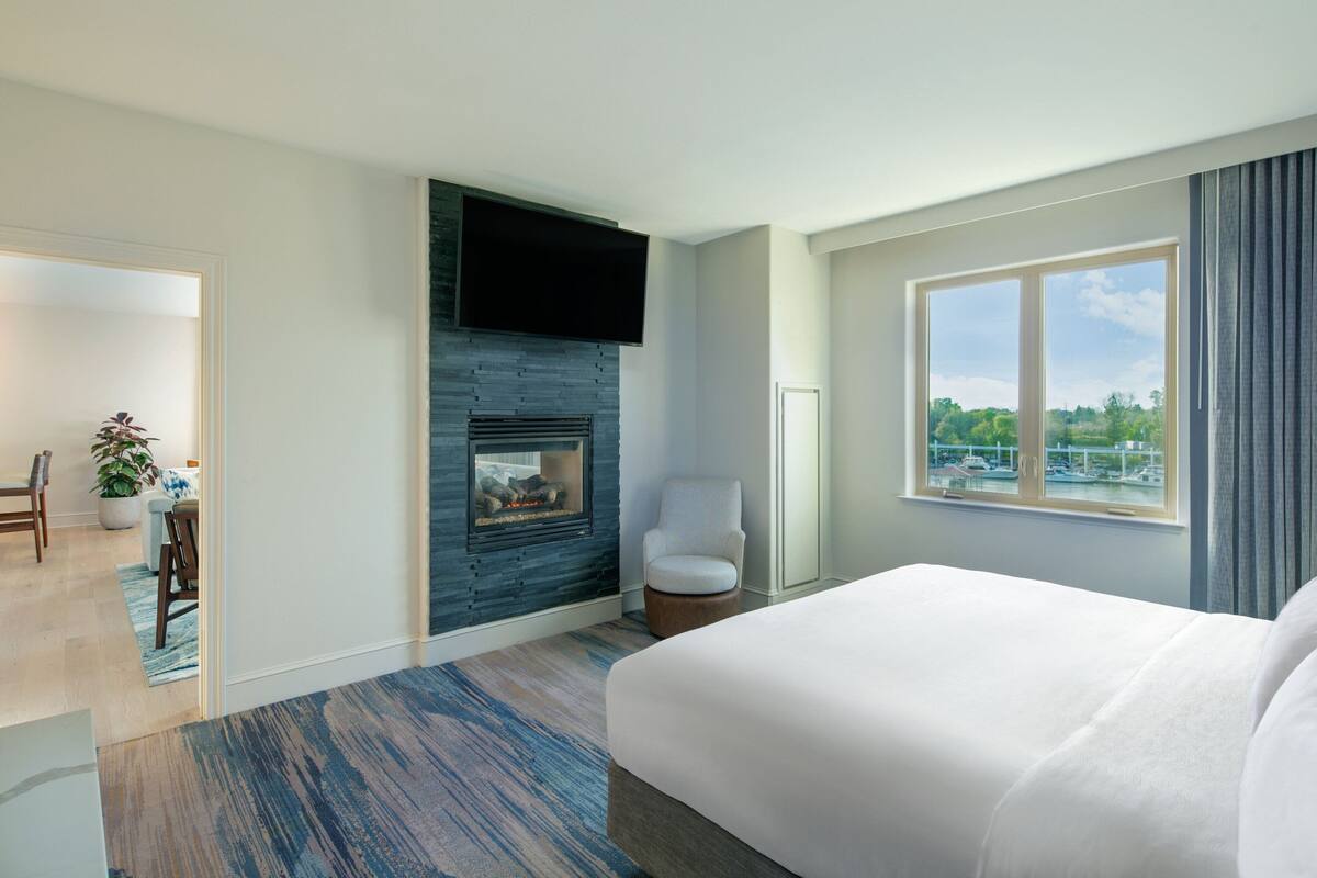 Una habitación de hotel moderna con una cama grande, una chimenea, un televisor montado en la pared y una ventana con vistas a la vegetación y barcos.