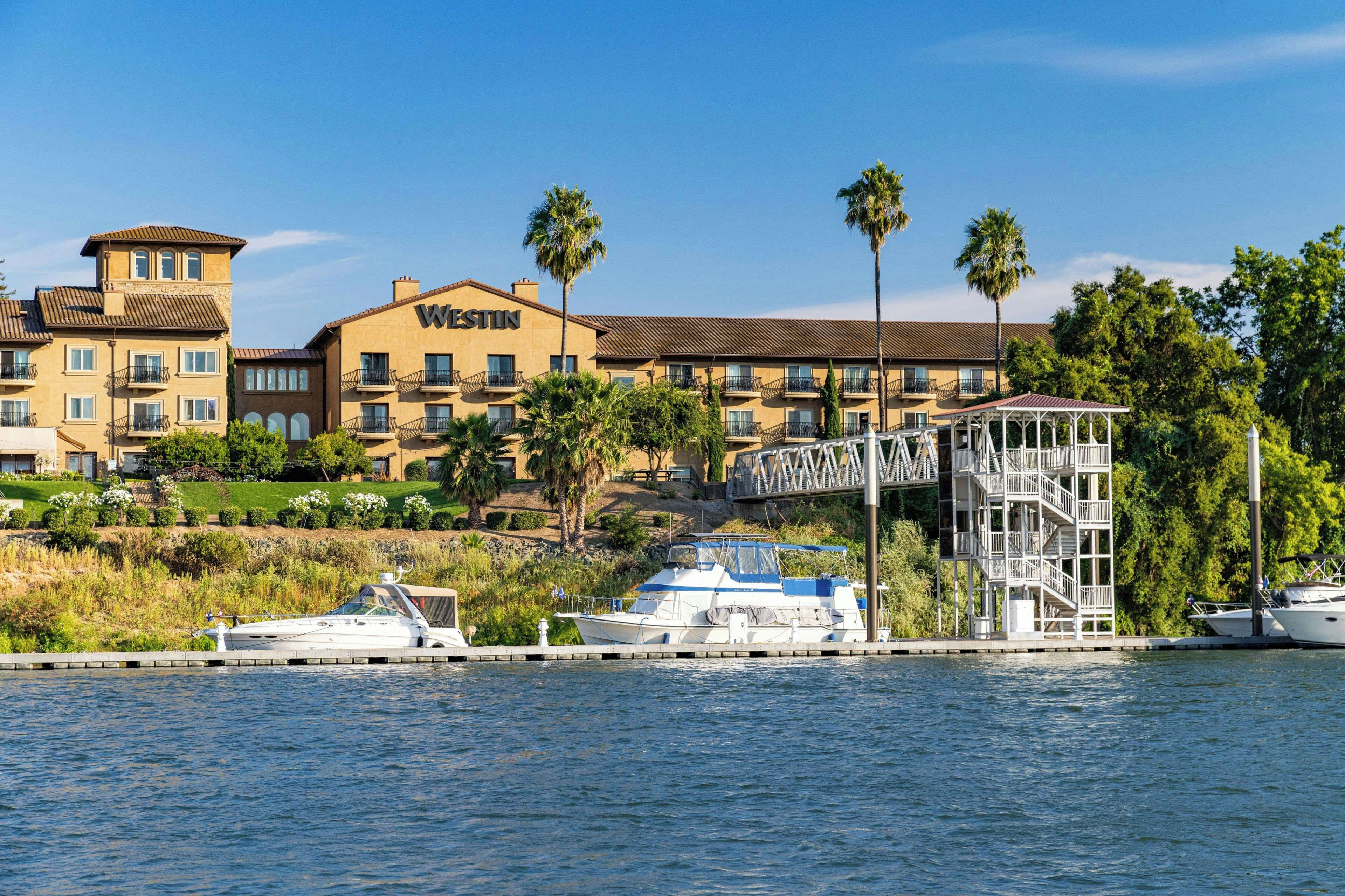 Un hotel frente al agua con varios balcones, un muelle con barcos y un edificio blanco con la señalización 'WESTIN'.