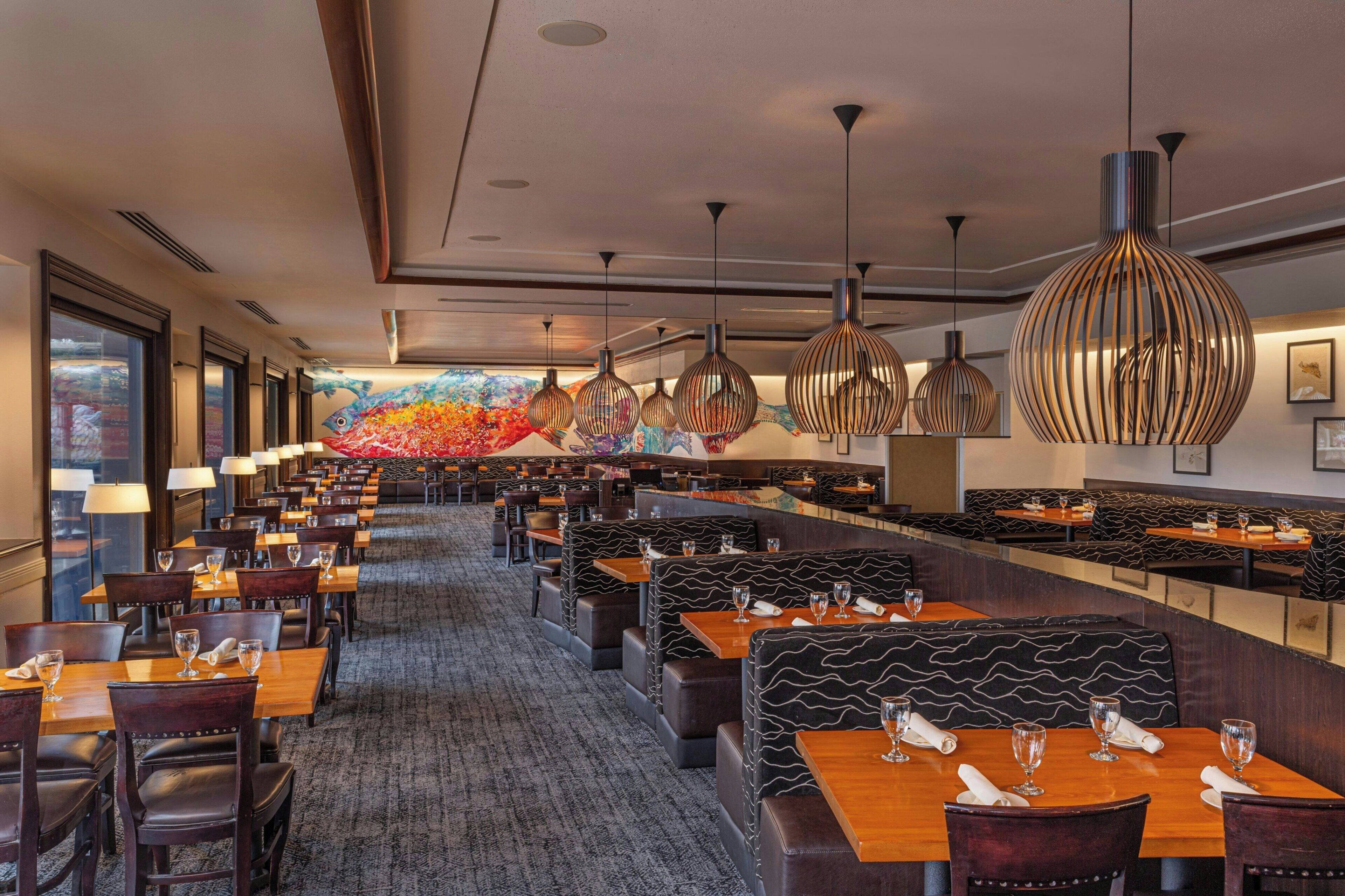 A restaurant with multiple dining areas, including booths and tables, with overhead lighting and a colorful mural on the back wall.