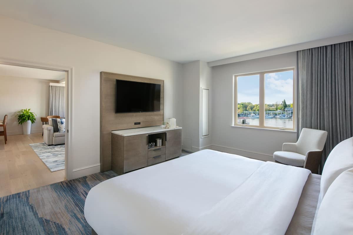 Habitación de hotel con una cama grande, un televisor de pantalla plana en la pared, una zona de estar con una silla y una mesita, y una ventana con vistas a un cuerpo de agua y edificios.