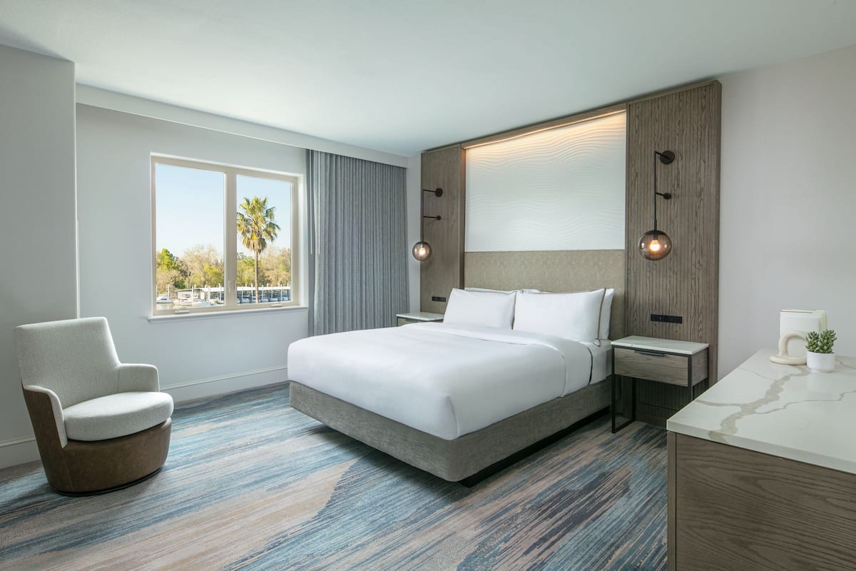 Una habitación de hotel moderna con una cama grande, una silla, un escritorio y vistas a palmeras a través de la ventana.