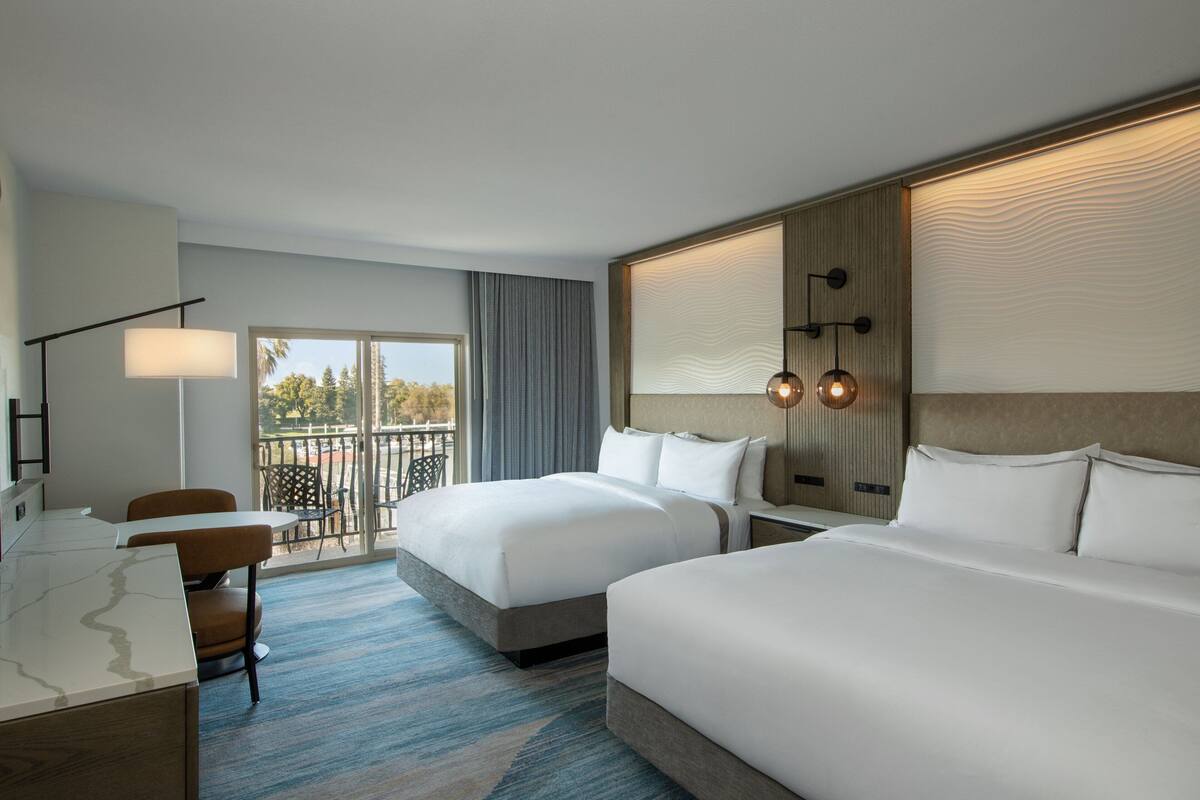 Habitación de hotel con dos camas, un escritorio, una silla y un balcón con vistas.