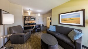 Une chambre d’hôtel moderne avec un canapé, un fauteuil et un coin repas, avec une cuisine en arrière-plan.
