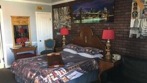 A hotel room with a bed, a desk, a chair, a mirror, a lamp, and a painting of a cityscape. - Travel Inn And Suites (Victorville)