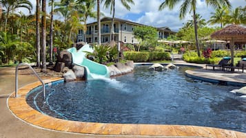 Relax by the resort pool surrounded by lush vegetation and palm trees, with a central water slide and lounge chairs nearby.