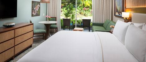 A hotel room with a large bed, a television, a small table with chairs, and a view of a garden through the window.