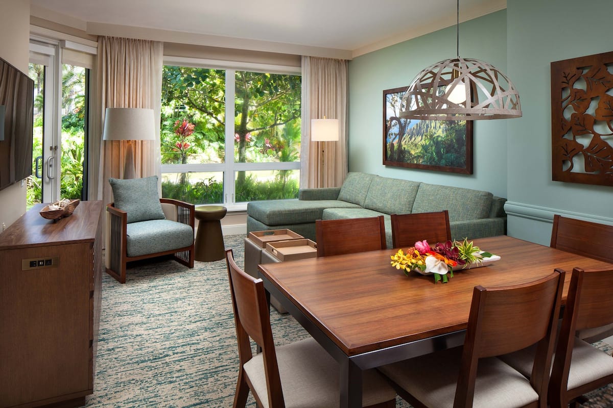 Habitación de hotel con zona de comedor, sofá, mesa de madera y un amplio ventanal con cortinas.