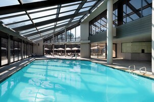 Una piscina con agua cristalina azul, rodeada de sillas de descanso y sombrillas, bajo una amplia estructura de techo de vidrio.