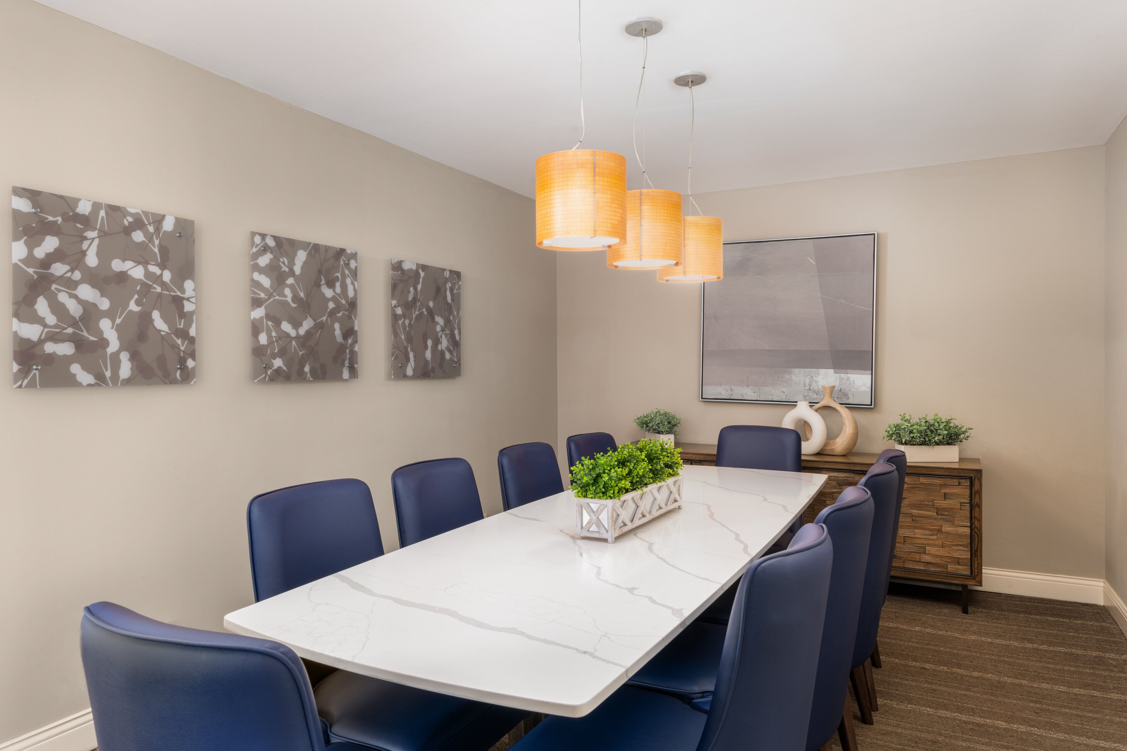 Une salle à manger moderne avec une table en marbre, des chaises bleues et des suspensions orange. Trois œuvres d’art abstraites ornent les murs, ainsi qu’un grand tableau encadré.