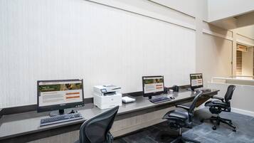 A modern office space with three computer workstations, each with a monitor, keyboard, and printer. There are two black office chairs and a wooden desk.