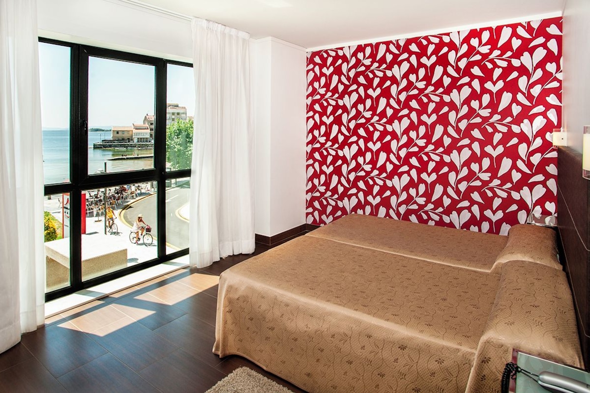 Habitación de hotel con un ventanal amplio que ofrece vistas al puerto y edificios, una cama con colcha estampada y una pared decorada con un diseño de hojas rojas y blancas.